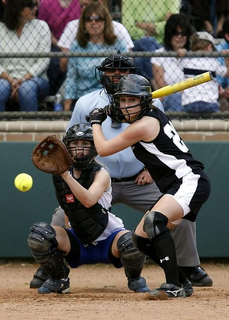 Youth softball batter in black swings at a pitch while catcher and umpire crouch behind home plate