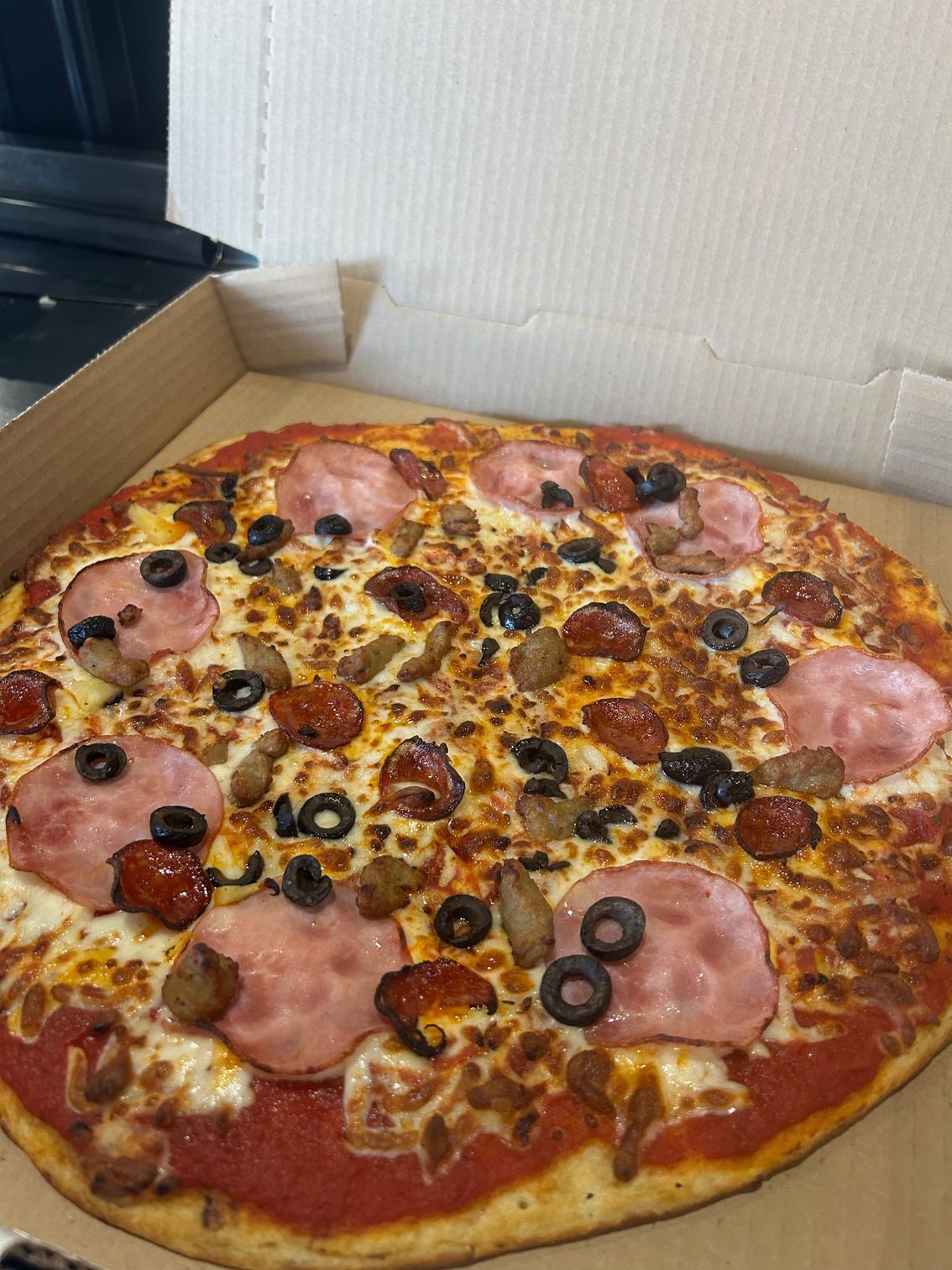 A pizza with ham , pepperoni , olives and cheese is in a box.