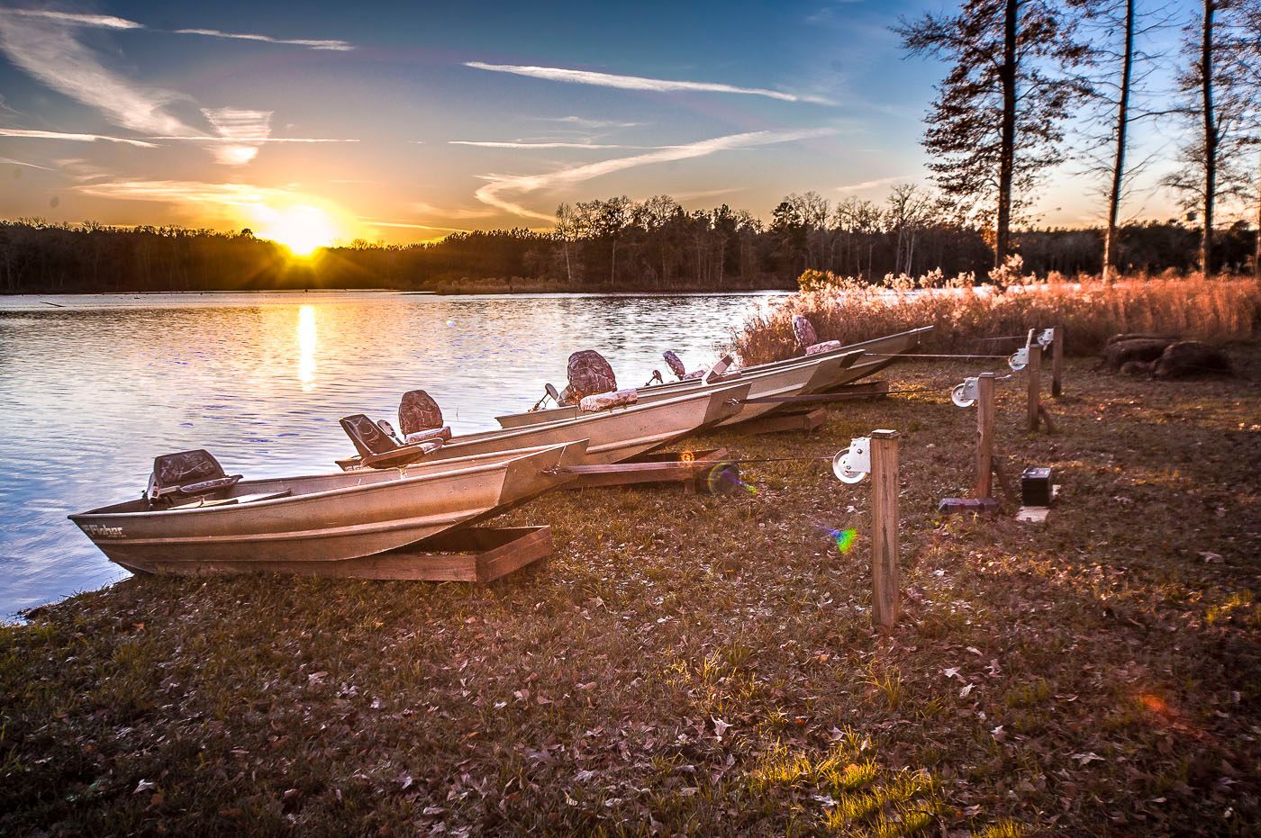 A row of boats are sitting on the shore of a lake at sunset.