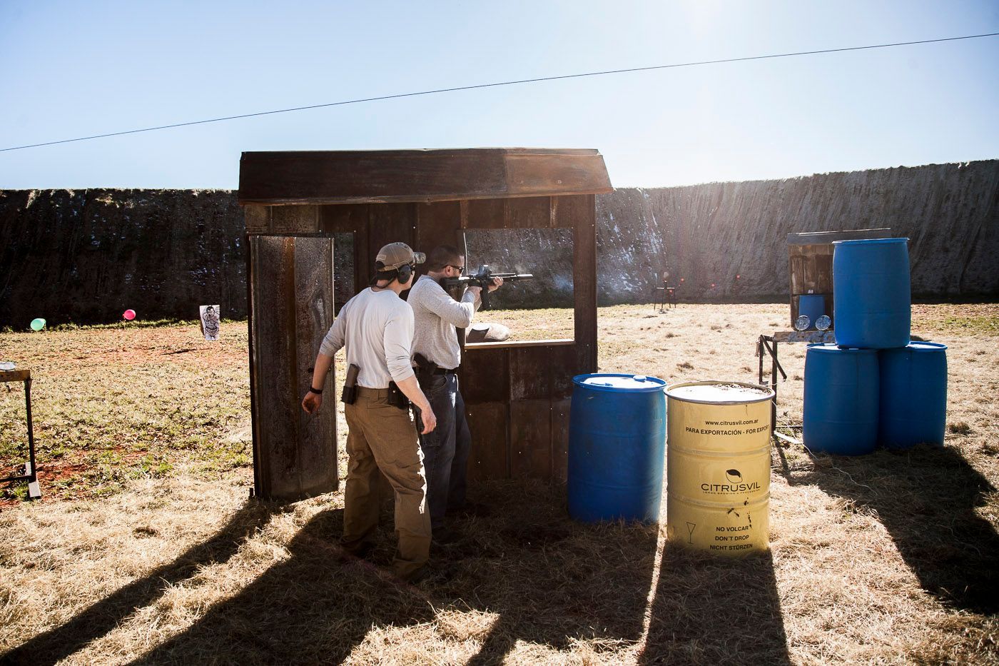 Two men are standing in a field with barrels in front of them.