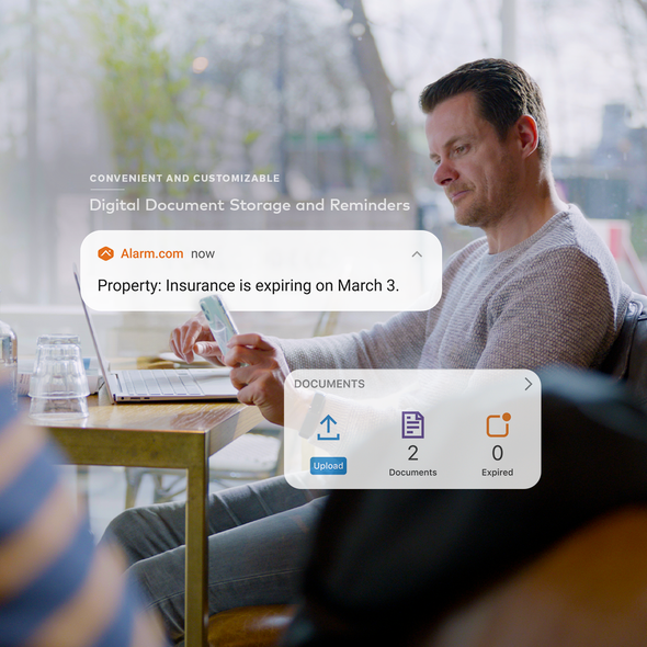 A man in a cafe uses his phone, which displays an Alarm.com insurance reminder and document storage interface.