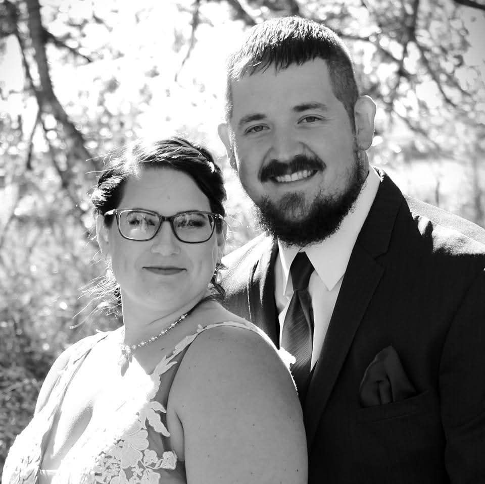 A black and white portrait of a smiling couple posing outdoors, with the woman wearing glasses and a lacy dress.