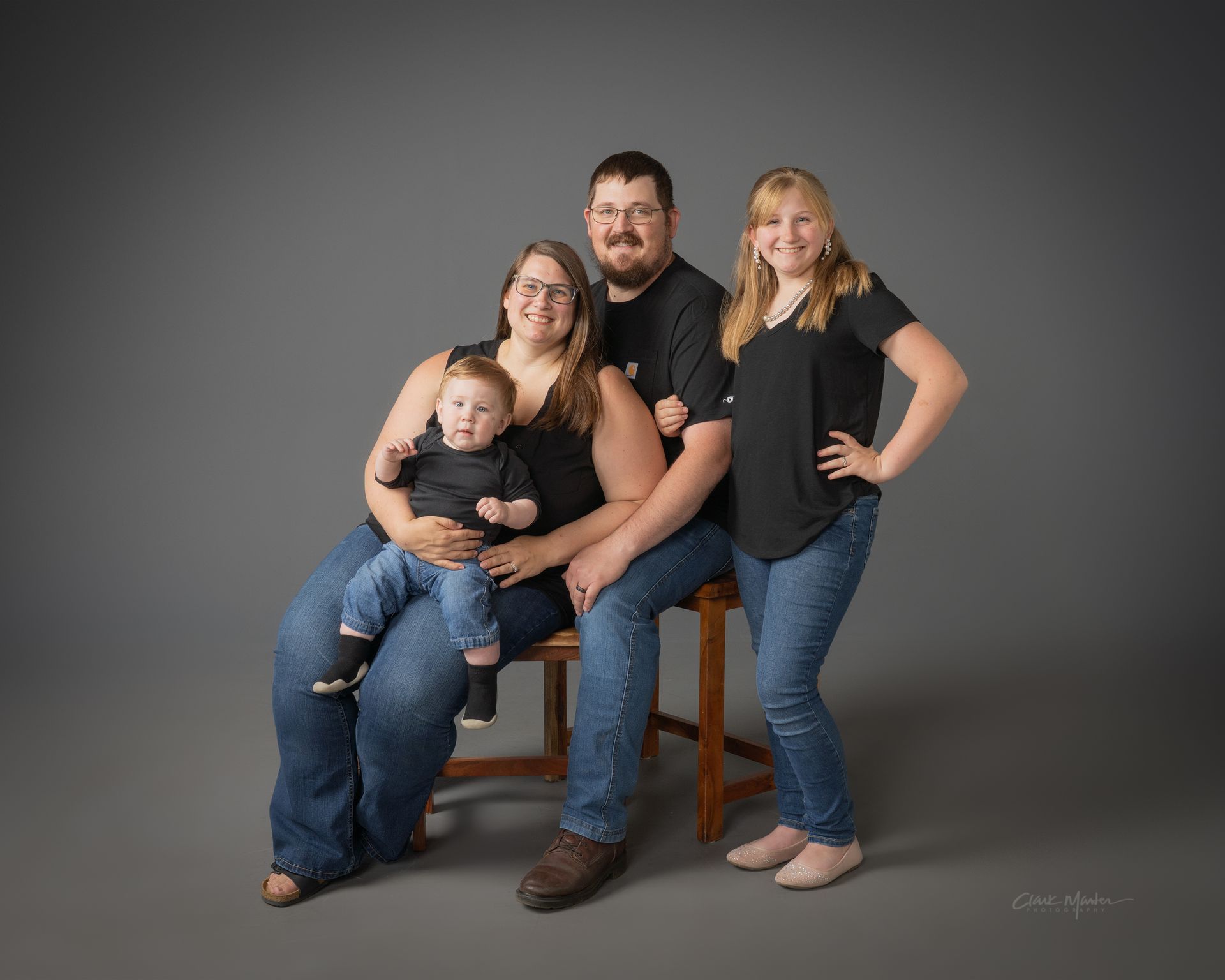 A family poses together in front of a gray backdrop, with two adults, a young person, and a toddler sitting on a lap.