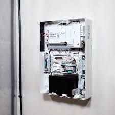 An open white alarm system control panel mounted on a wall, showing internal circuit boards, wiring, and a black battery.