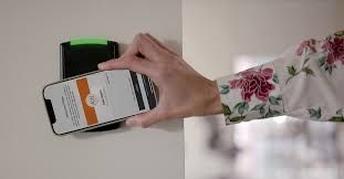 A person holds a smartphone near a wall-mounted electronic card reader to scan a digital access credential.