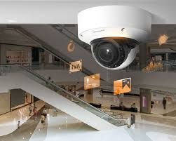 A ceiling-mounted security camera overlooks a modern, multi-level indoor shopping mall with escalators and people.