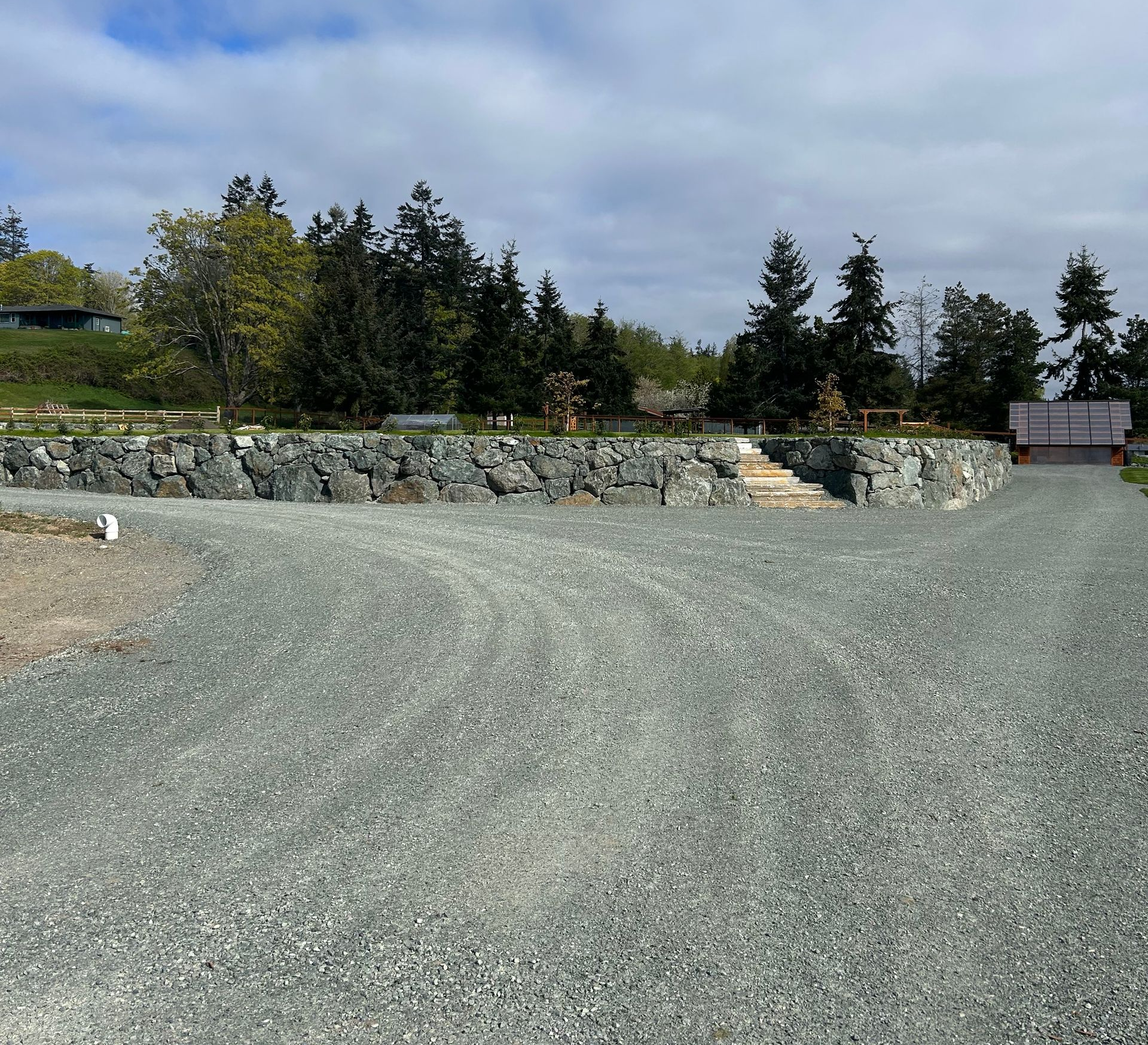 Gravel parking area in front of a stone retaining wall with steps, leading to trees under a cloudy sky.