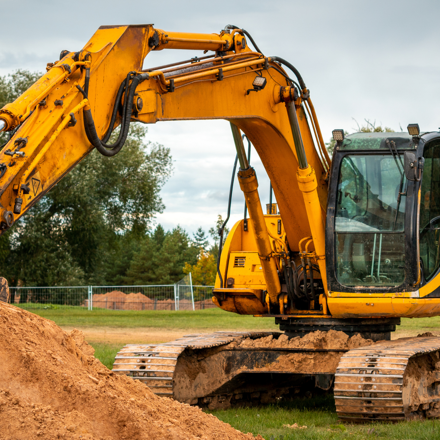Yellow excavator digging in a grassy area, small pile of dirt in front. Overcast sky.