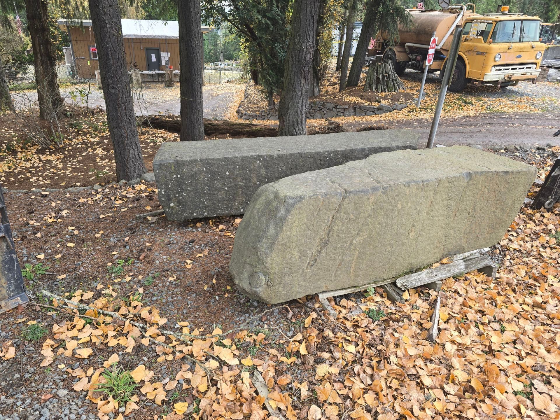 Forklift parked near stone blocks and a stone wall in a wooded area with power lines in the background.