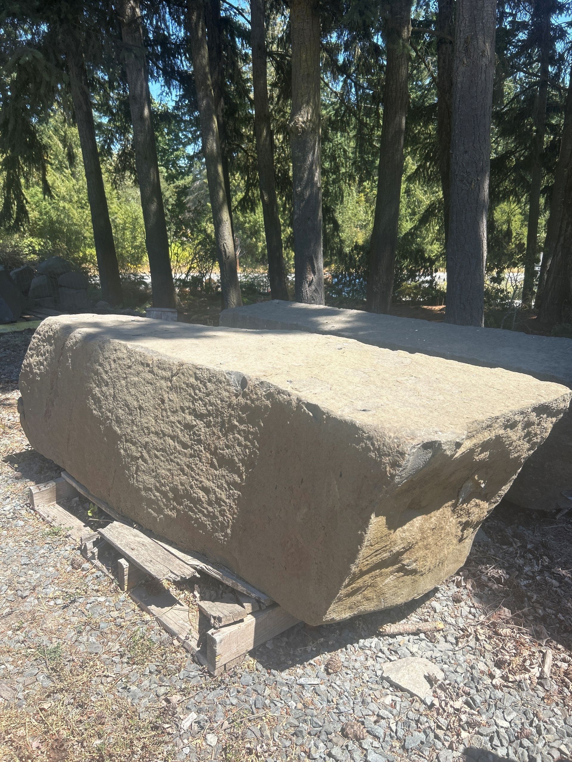 Large, rectangular stone block outdoors, sitting on a base. Trees in the background, sunny day.