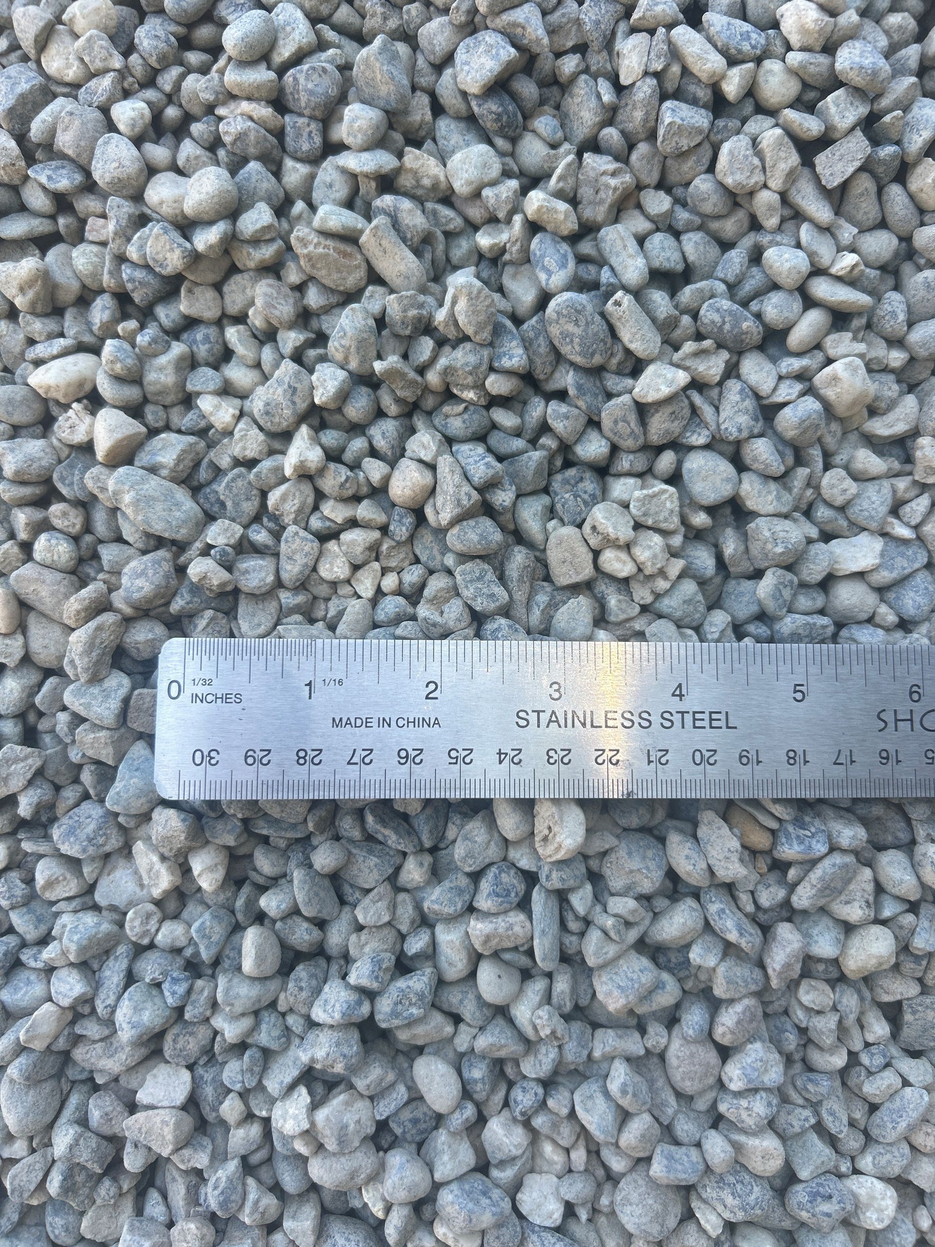 Close-up of small gray gravel rocks with a stainless steel ruler for scale.