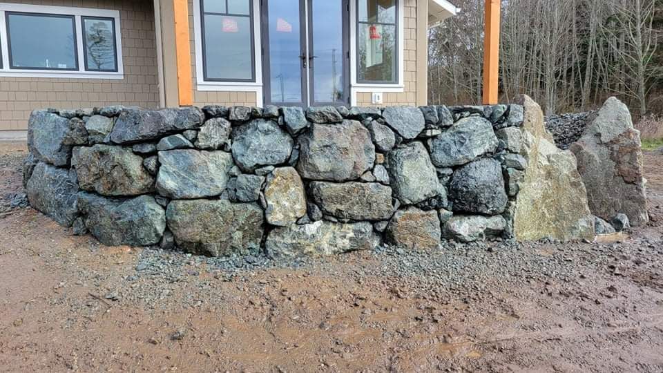 Stone retaining wall in front of a house with gray and blue rocks. Brown dirt and gravel in foreground.