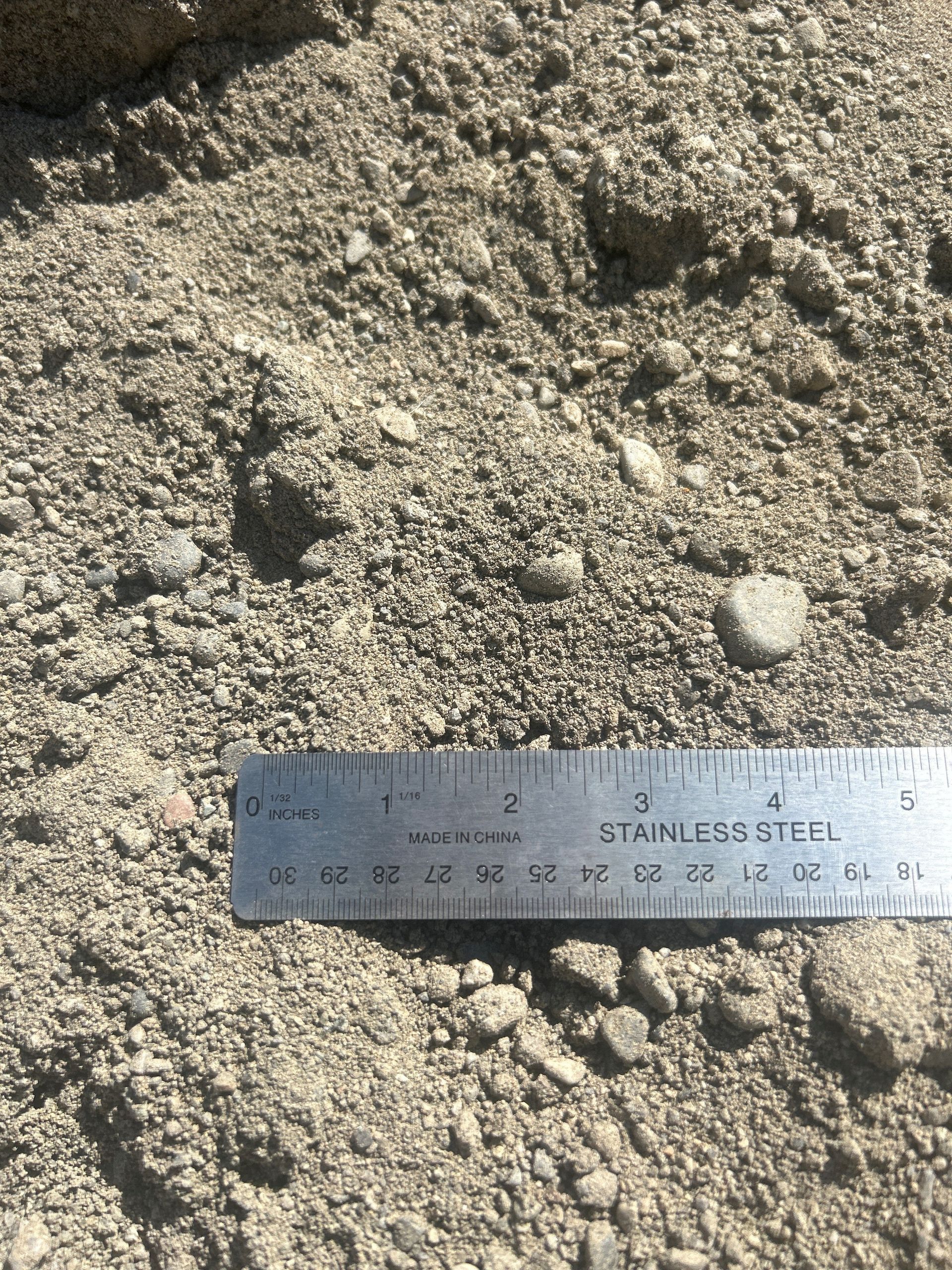 Close-up of gravel, sand, and small rocks with a stainless steel ruler, showing size comparison.