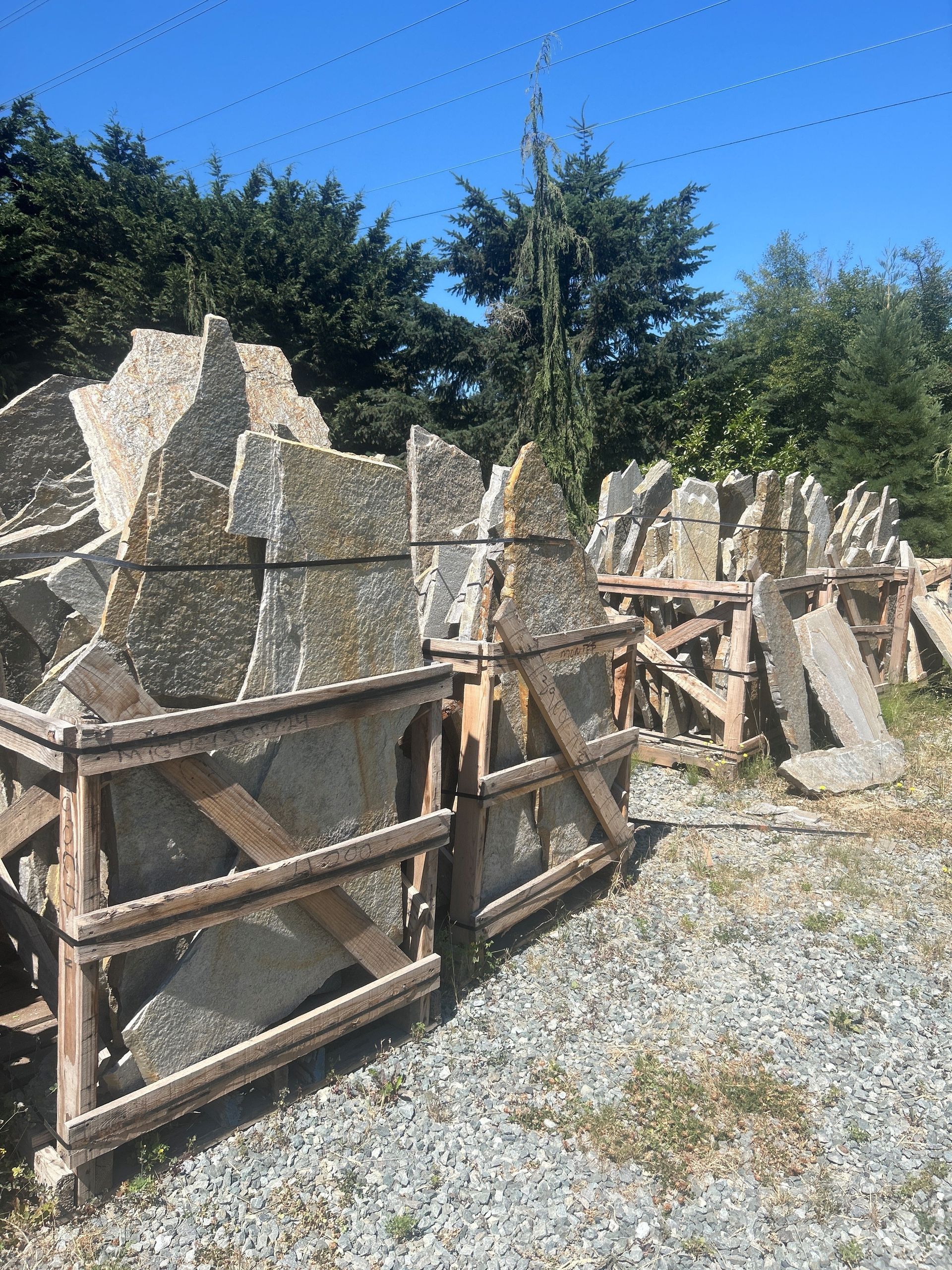 Stone slabs stacked in wooden crates, outdoors. Green trees and blue sky in background.