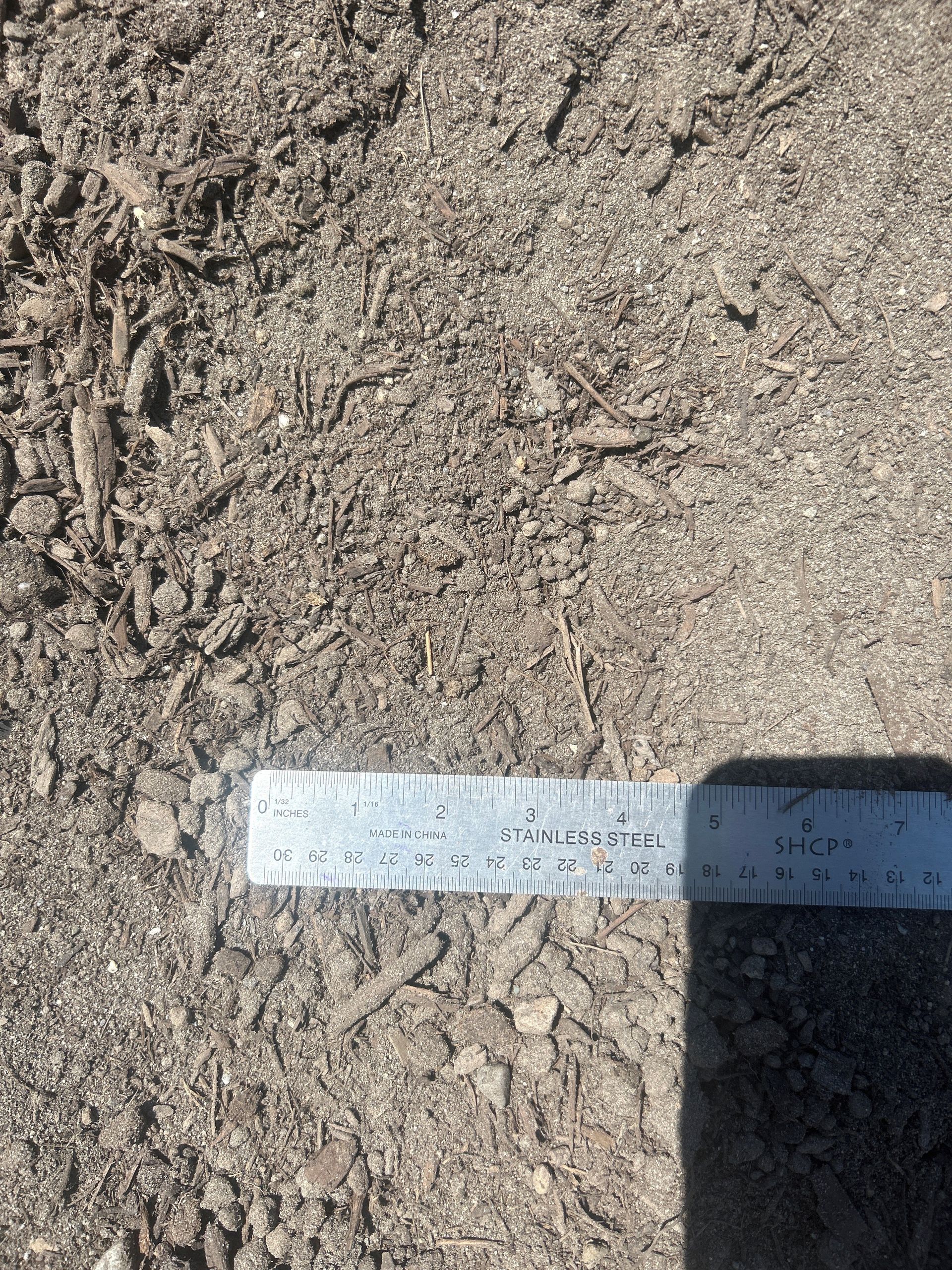 Close-up of brown soil with a metal ruler for scale, showing various textures and small debris.