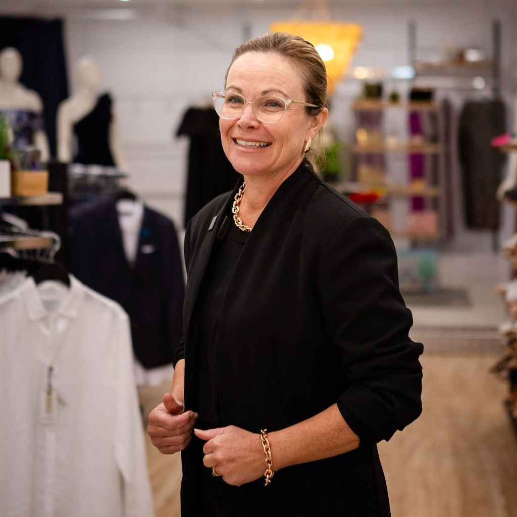 A woman wearing glasses and a black jacket is smiling in a clothing store.