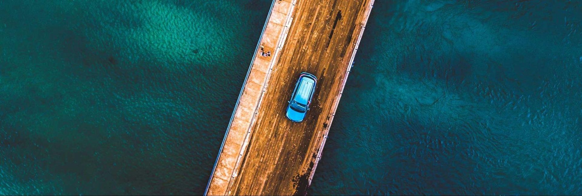 An aerial view of a car driving on a wooden bridge over a body of water.