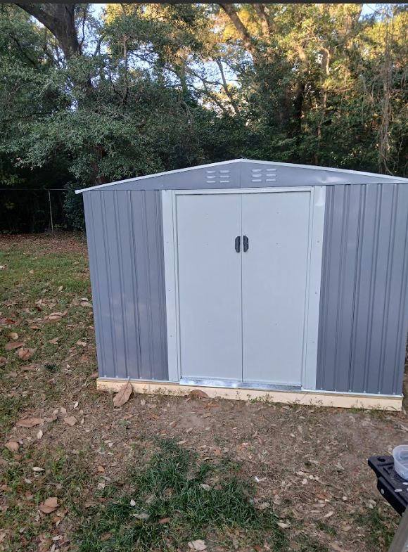 Gray metal shed with light gray double doors in a yard, surrounded by trees and grass.