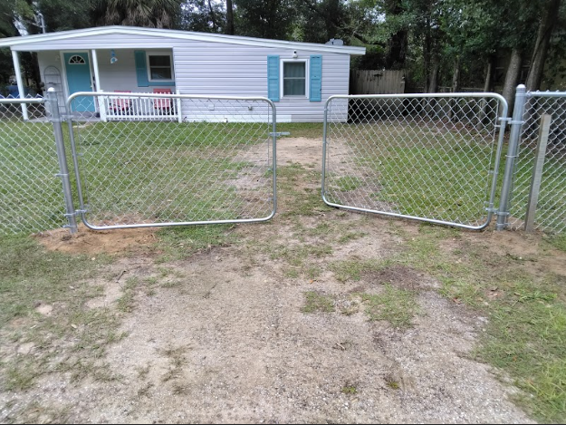 Chain-link fence with open gates leading to a light blue mobile home. Green grass and dirt driveway.