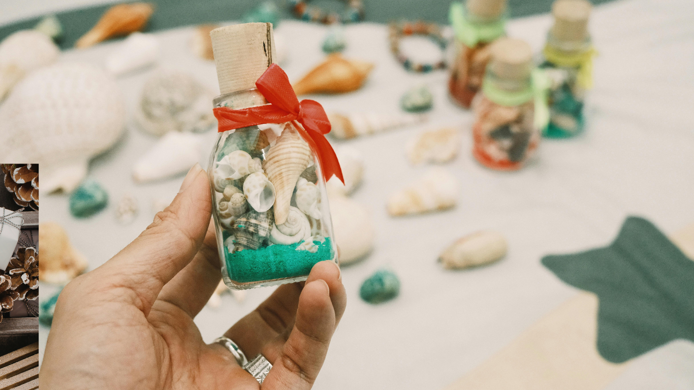 Hand holding a small bottle filled with seashells, red ribbon tied on top, surrounded by other seashells.