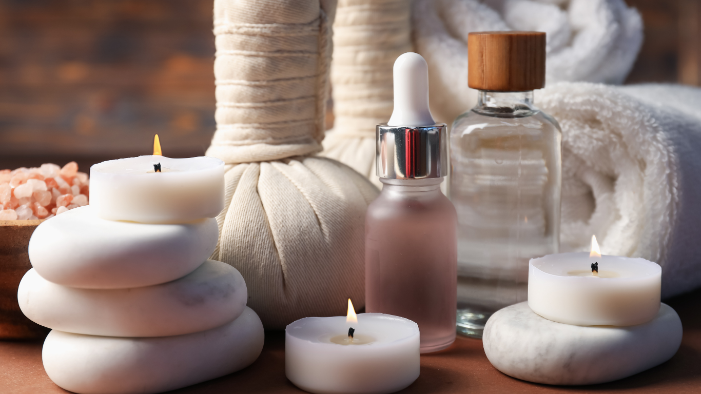 Spa setting with lit candles, massage stones, oil bottle, towel, and salt.
