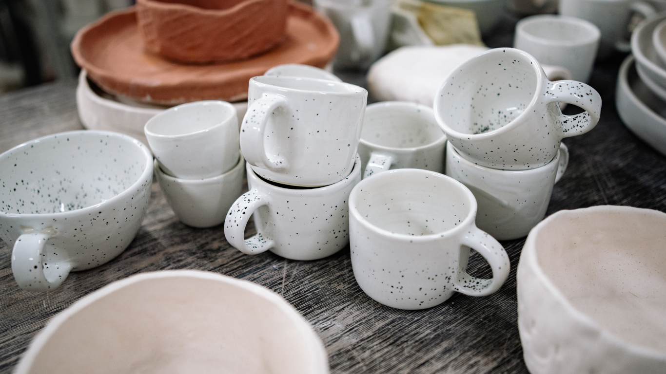 Assortment of handmade speckled white ceramic mugs and bowls on a textured surface.