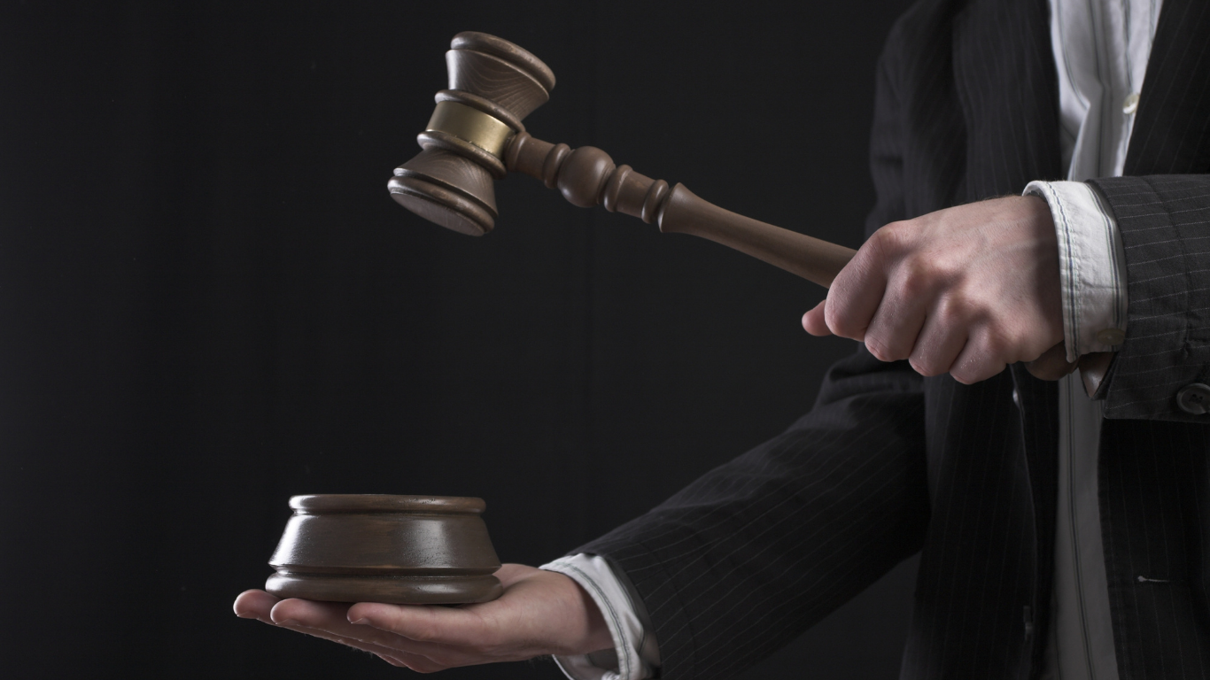 Hand holding a gavel, about to strike a wooden block. Dark background.