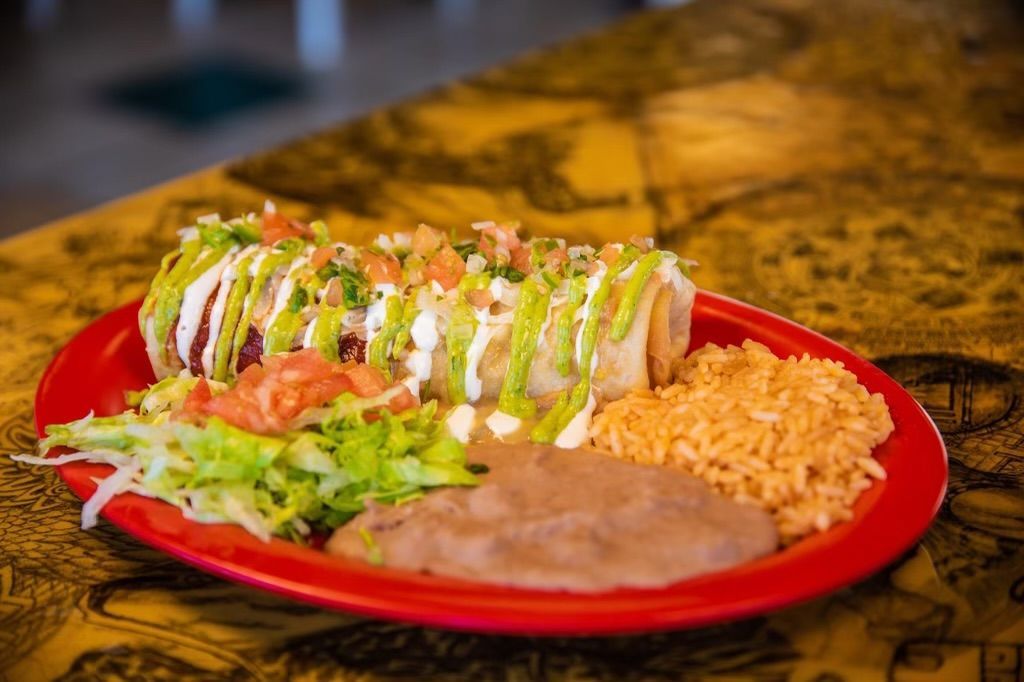A red plate topped with a burrito , rice and beans on a table.
