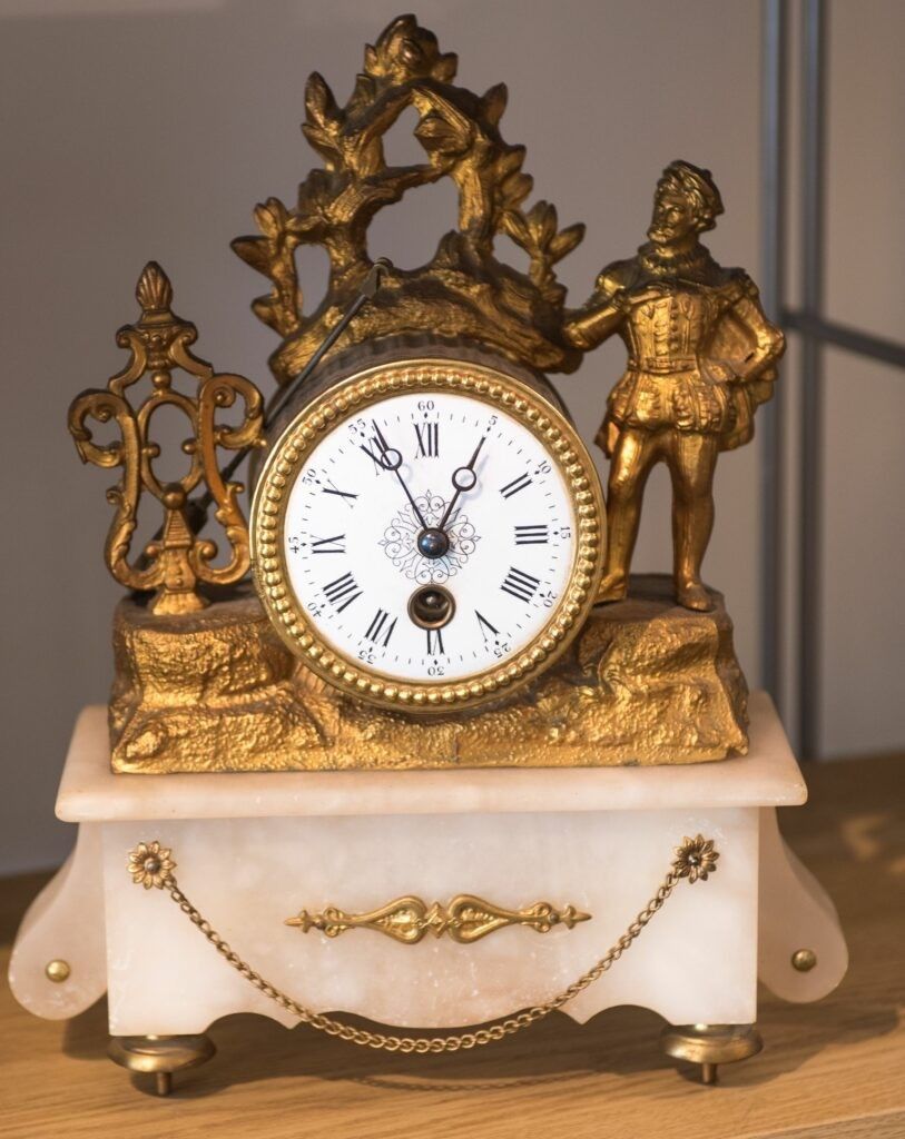 Gold-toned antique clock with a figure on top and ornate details, set on a white marble base.