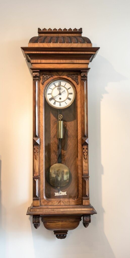 Wooden wall clock with pendulum and ornate carving, against a cream-colored wall.