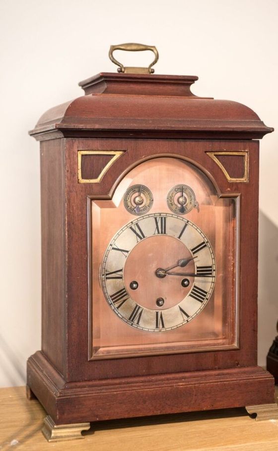 Wooden antique clock with a copper face, Roman numerals, and brass handle on top.