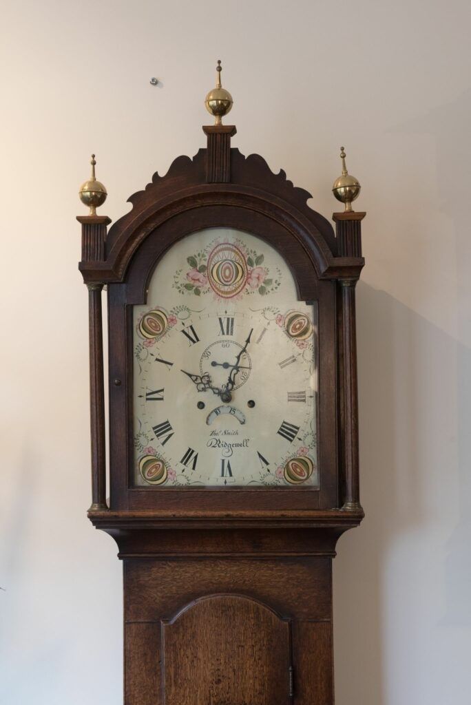Tall, wooden grandfather clock with a white face, Roman numerals, and floral accents.