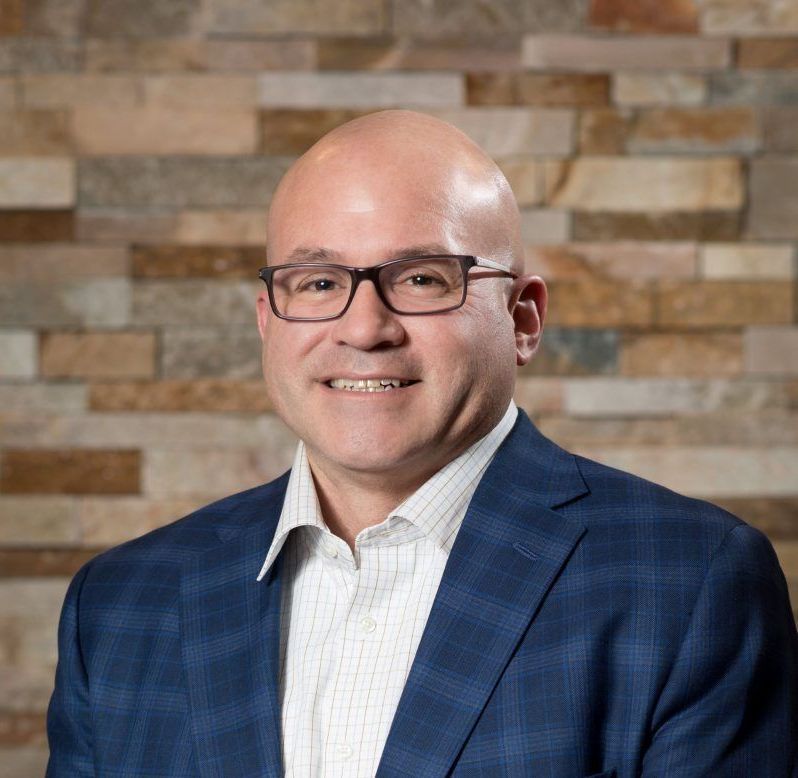 Bald man in glasses and blue plaid blazer smiles in front of a stone wall.