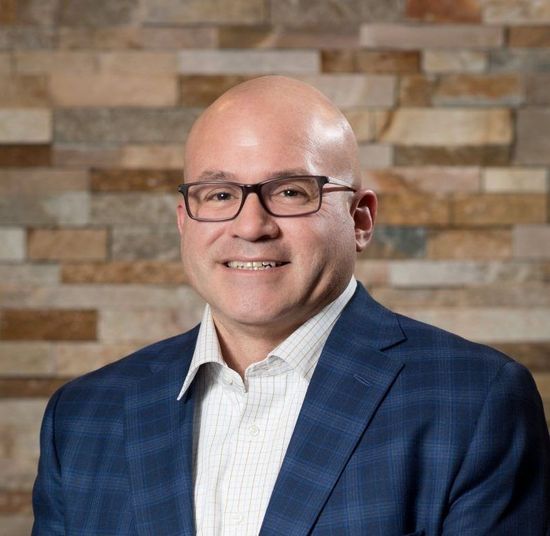 Bald man in glasses, blue plaid blazer, smiles in front of a textured stone wall.