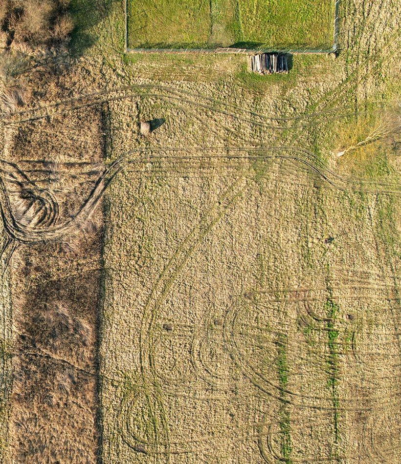 Aerial view of a field with tire tracks, hay bale, small shed, and fenced-in grassy area.