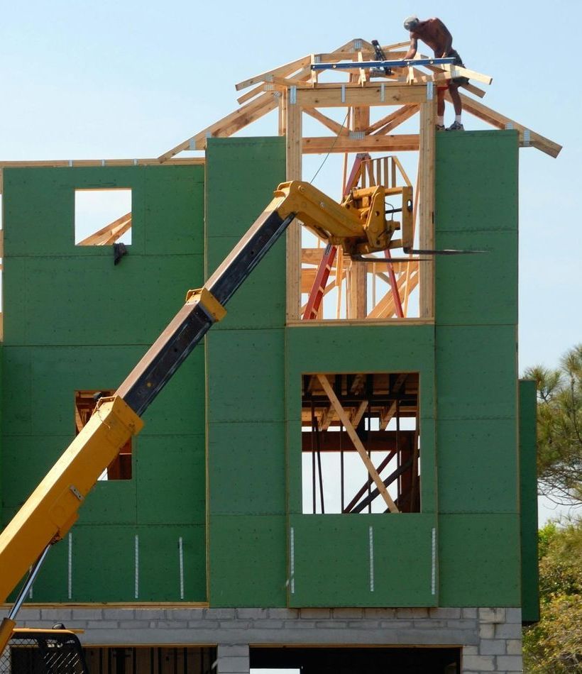 Construction worker on roof frame; a crane lifts materials to the second story, green siding. Sunny, outdoor setting.