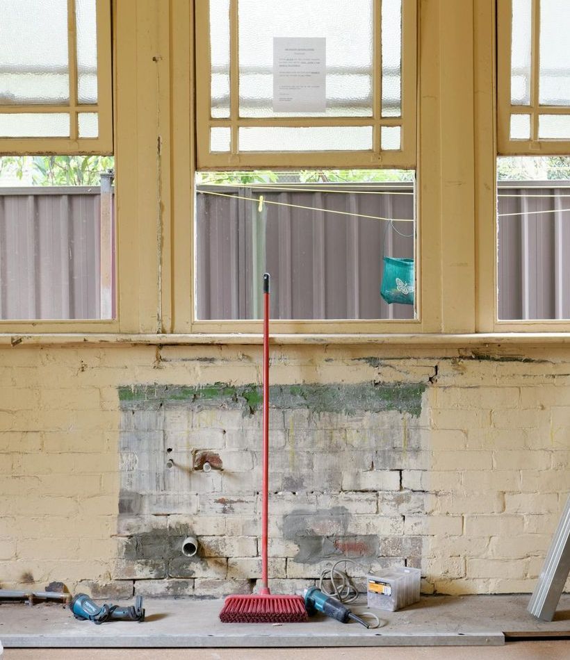 Interior view of a room under renovation; brick wall with exposed patches; red broom stands against the wall.
