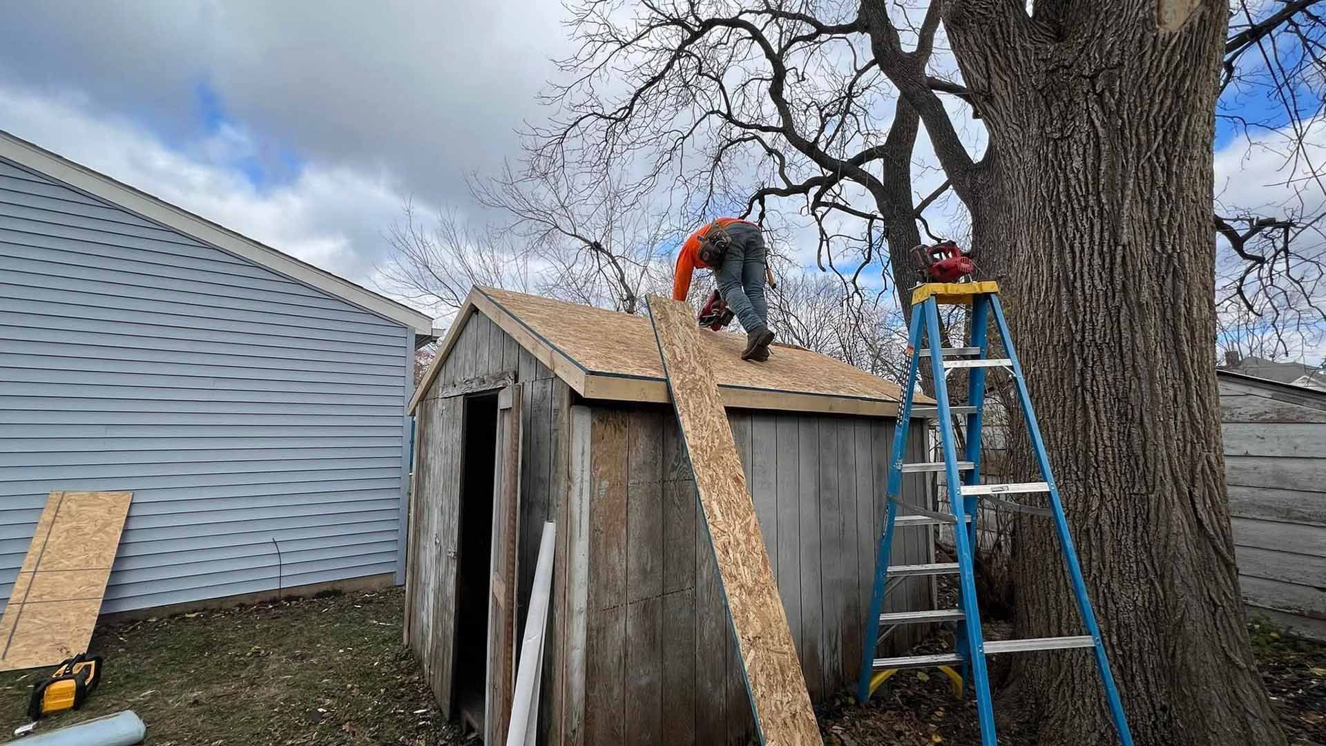 A man on a ladder is working on the roof of a shed