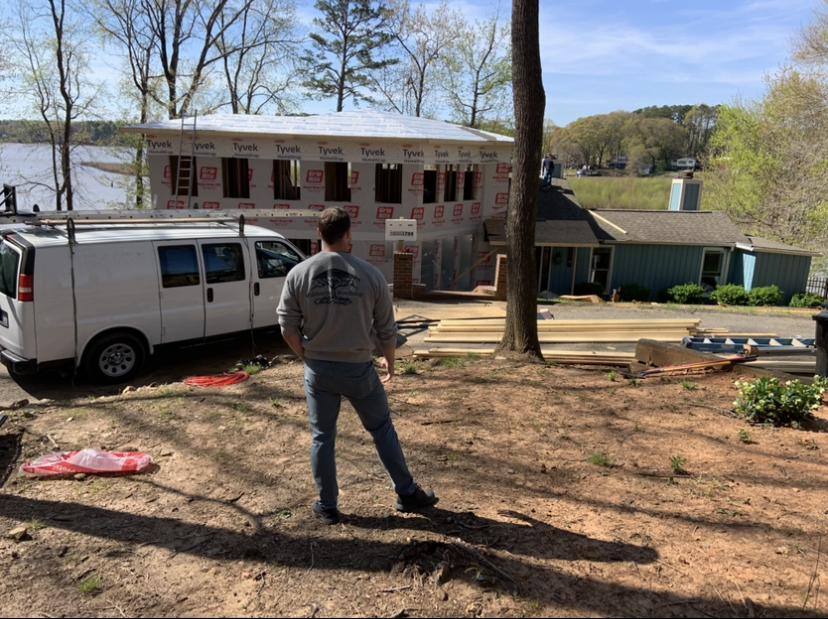 Man overlooking a house under construction next to an existing blue house and a white van. Sunny day.