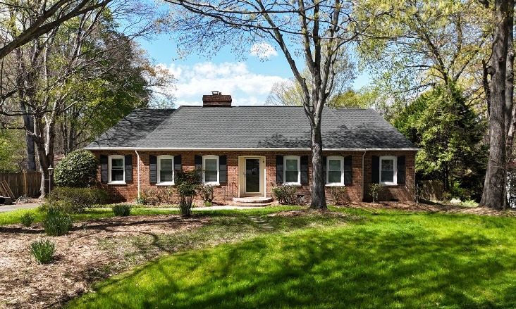 Brick ranch house with black shutters and a green lawn surrounded by trees.