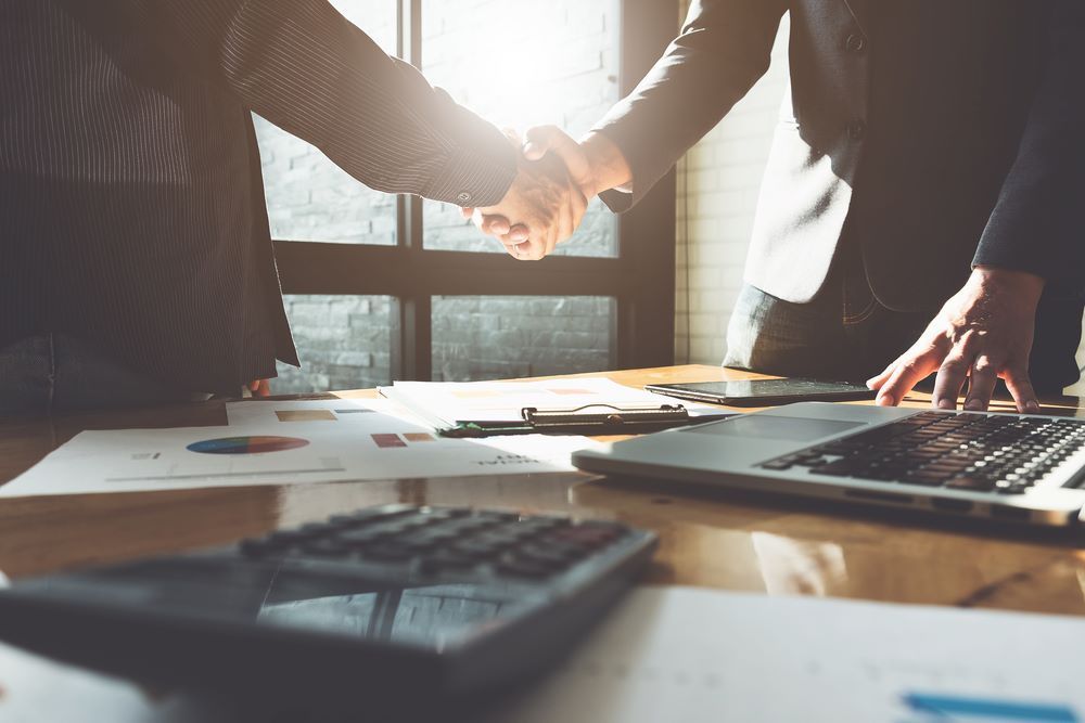 Two Businessmen Are Shaking Hands Over a Table With a Laptop and Calculator — D&T Accounting & Bookkeeping In Gosford, NSW