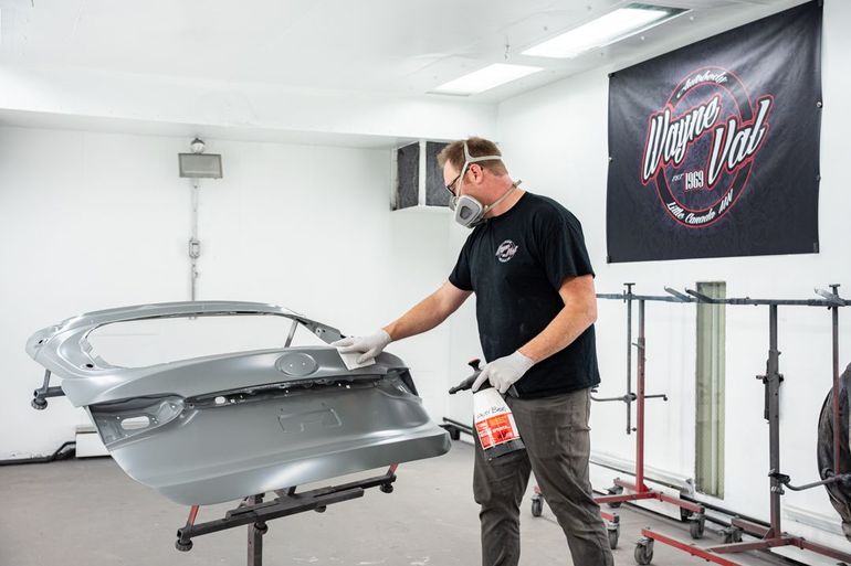 Man in respirator prepares car trunk for paint in a bright workshop.
