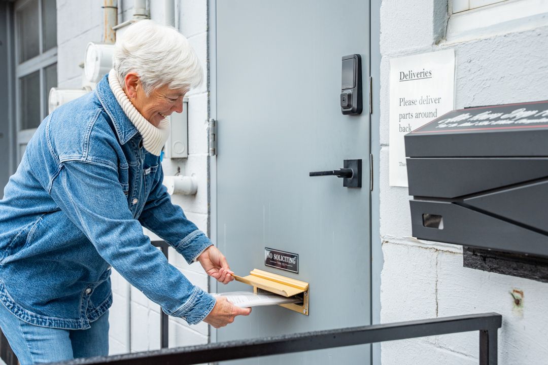 Woman with white hair in denim jacket retrieving mail from a door slot.