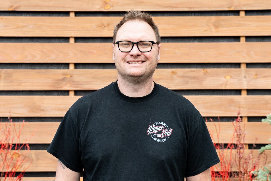 Man wearing glasses and a black t-shirt smiles in front of a wood fence.