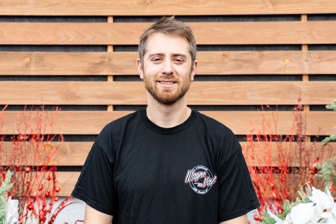Man with a beard in a black shirt smiles in front of a wooden wall with red accents.