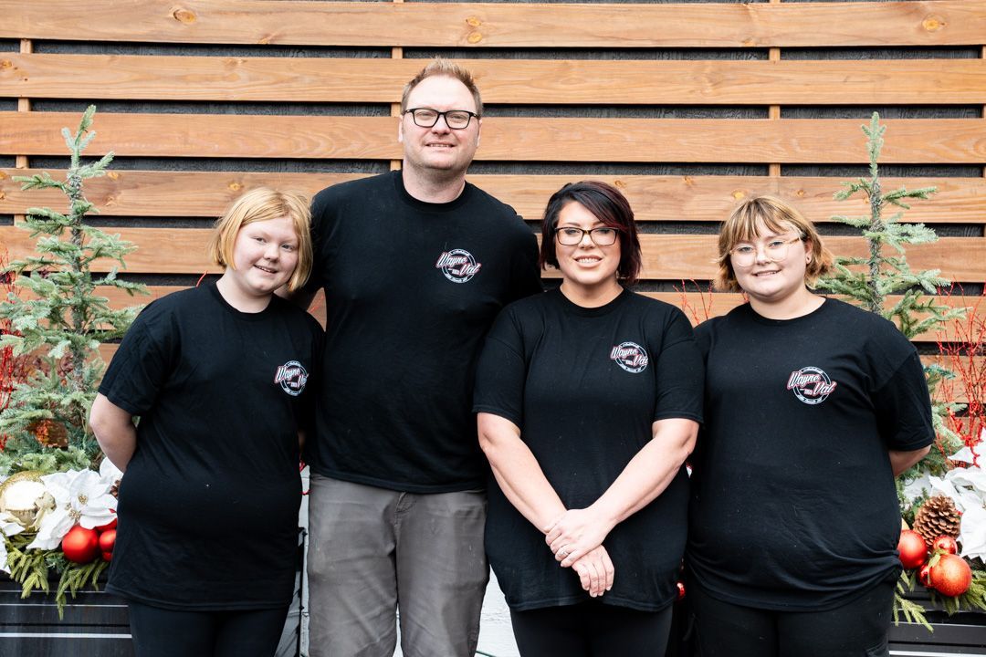 Four people wearing black shirts pose outdoors in front of a wood background.