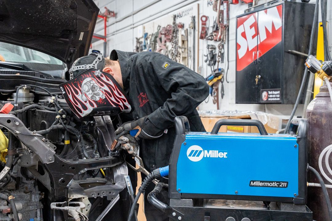 Welder wearing mask, working on a car in a shop, blue Miller machine in foreground.