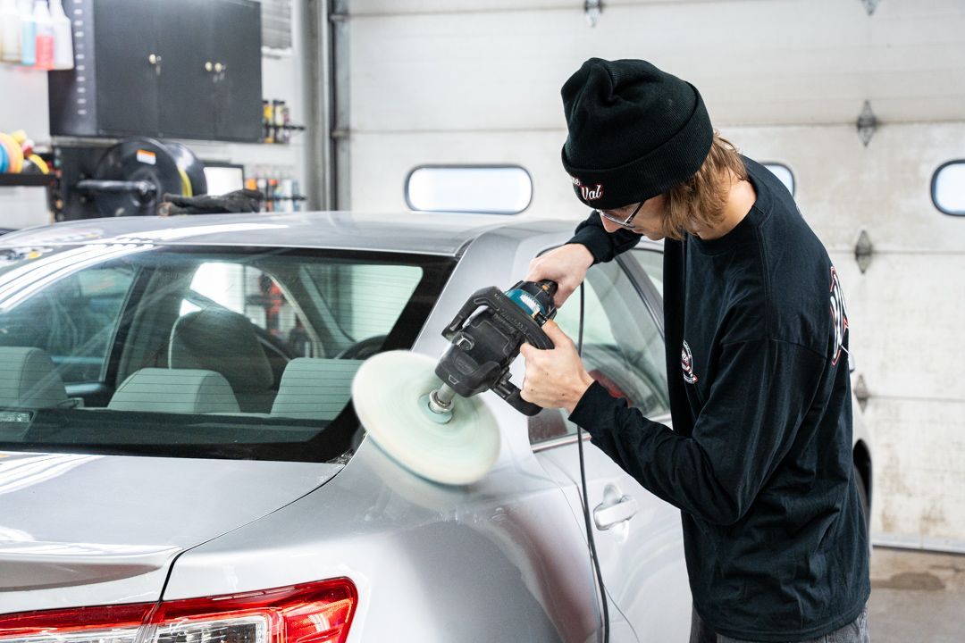 A person polishing a silver car in a garage with a power buffer.