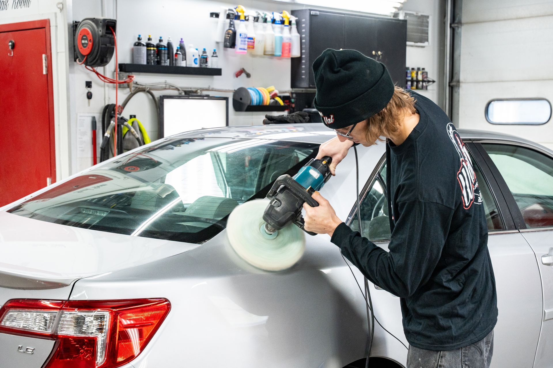 Person polishing the side of a silver car with an electric buffer in a garage.