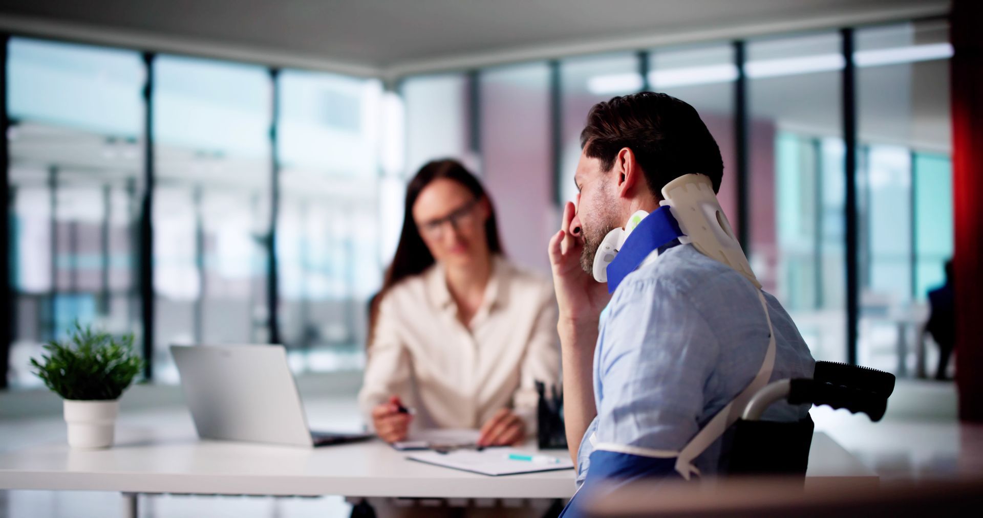 Two coworkers in a modern office meeting, one talking while the other listens across a desk.